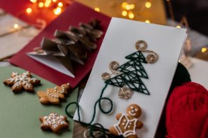 Handmade craft paper Christmas New Year card with wool thread embroidering. Concept of holiday handicraft at home, family quality time with children. Lights, gingerbread cookies on wooden background